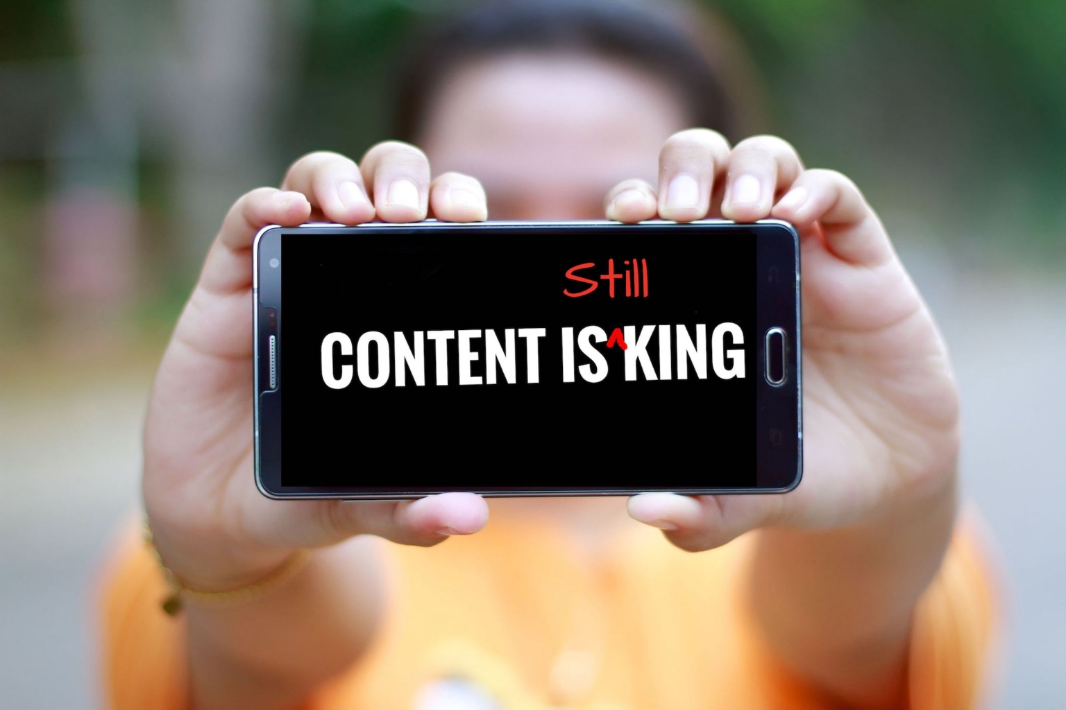 A woman in yellow holds up a cellphone with 'Content is King' on the screen. In red, the word "still" has been added, so that the text reads "content is still king"